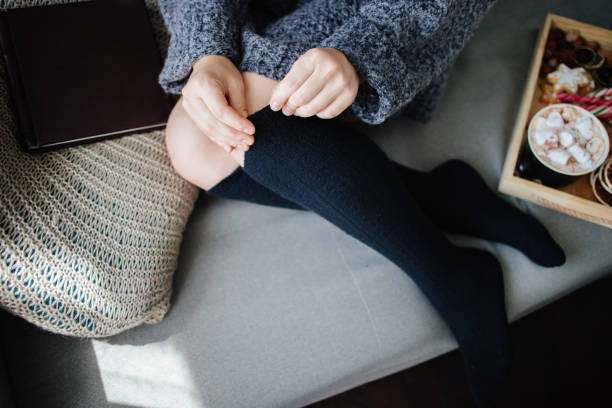 Woman on couch pulling up cozy black compression socks for women, enjoying a warm drink.